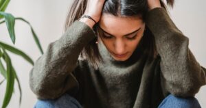 Woman covering her face with both hands, wearing a cozy sweater, seemingly overwhelmed or distressed.