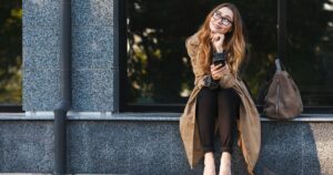 Young woman with glasses sitting on a city ledge, smiling and holding a phone, lost in thought. A brown handbag sits beside her.