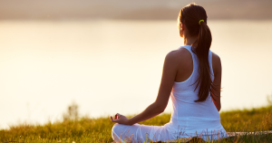 Woman in white sitting cross-legged outdoors at sunset, practicing meditation peacefully near water.