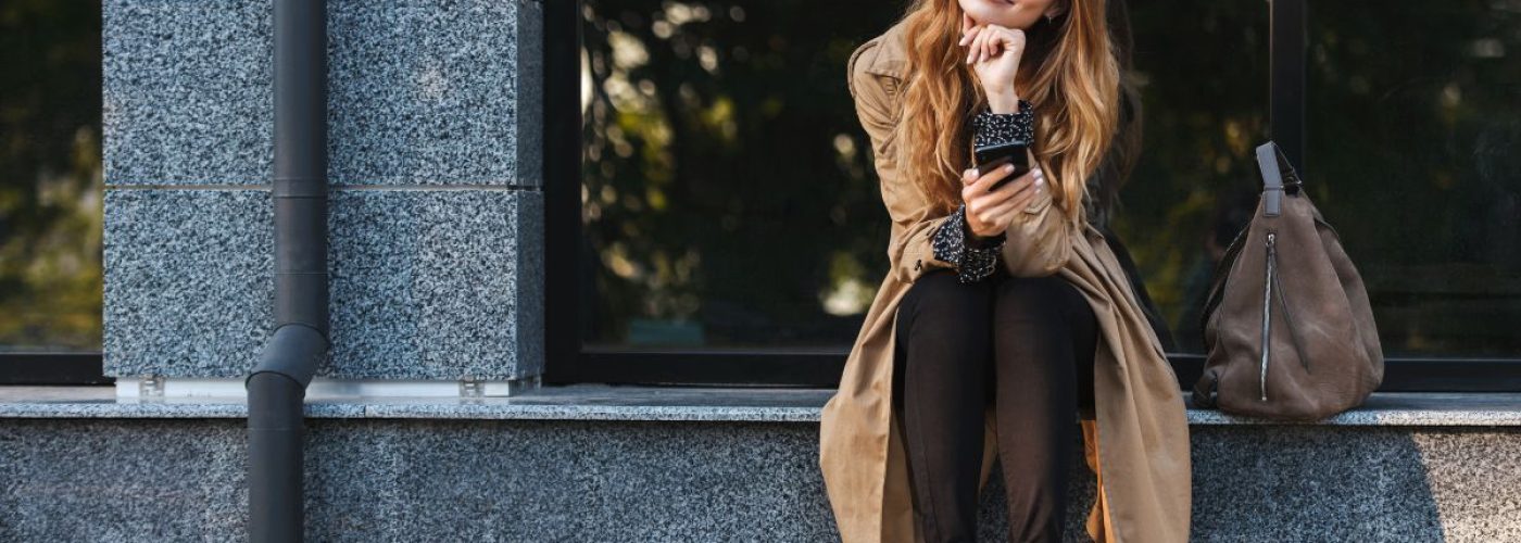 Young woman with glasses sitting on a city ledge, smiling and holding a phone, lost in thought. A brown handbag sits beside her.