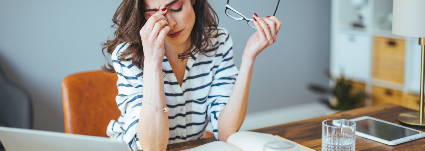 A stressed woman sitting at her desk holding her head, showing signs of burnout and anxiety while working on her laptop.